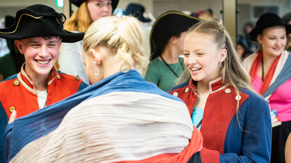 Two girls and a boy aged about fourteen are trying on costumes from Les Miserables. Two are wearing red and blue tunics, and another has a blue, white and red French flag wrapped around themselves. The boy also wears a black hat.