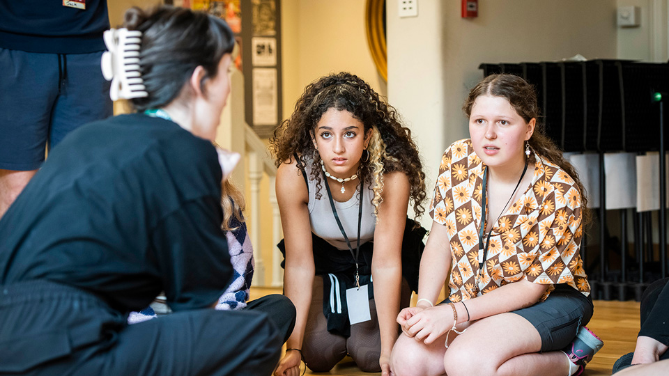 A female workshop leader dressed in black is crouching in front of two kneeling girls aged about sixteen. The girls are listening intently to what the workshop leader is telling them.