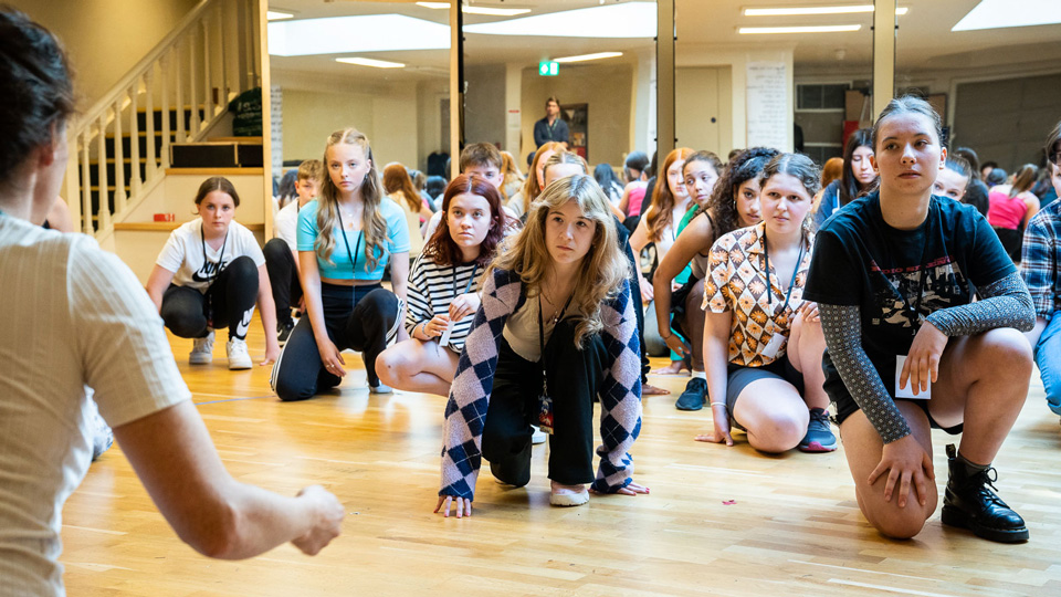 A group of students crouching on the floor listening to a workshop leader giving them instructions. Everyone is dressed in casual clothing.