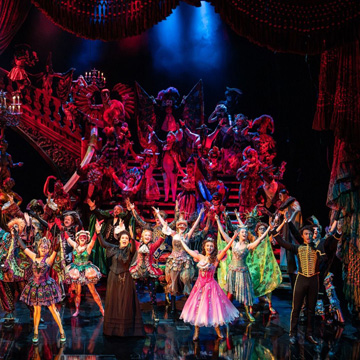 A photograph of the Masquerade number from The Phantom of the Opera. The full cast is stood around a large sweeping staircase, and everyone is dressed in colourful fancy dress costumes with their arms raised.